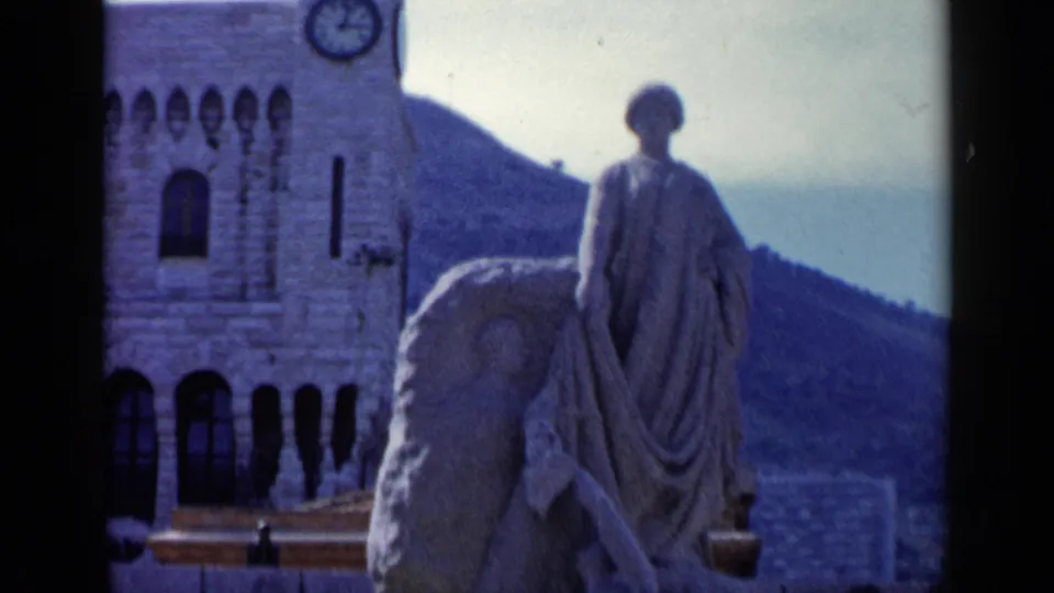 1960 8mm frame showing a draped stone statue in the foreground with a clock tower, arched building details, benches, and hillside beyond.