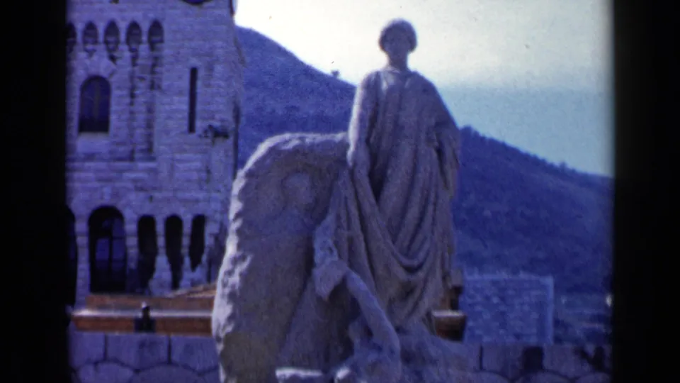 1960 8mm travel frame with a closer view of the draped stone statue, arched stone facade, hillside, and a small portion of distant city below.
