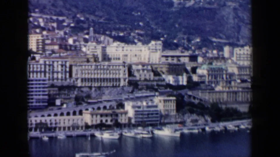 1960s 8mm frame of a coastal waterfront, showing moored boats, pale urban buildings, and a dense hillside backdrop.