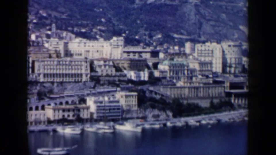 Soft 8mm travel frame showing a harbor-side urban district with boats in the foreground and apartment-like buildings on the slope beyond.