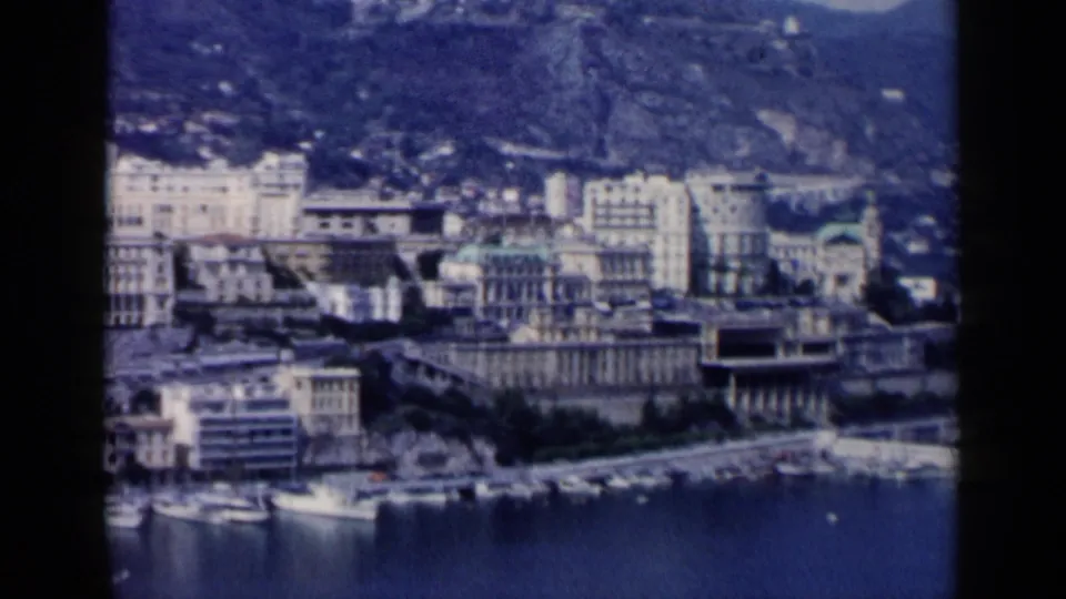 1960s 8mm frame of a European-looking harbor city, with marina boats below pale waterfront buildings and hillside development in the distance.