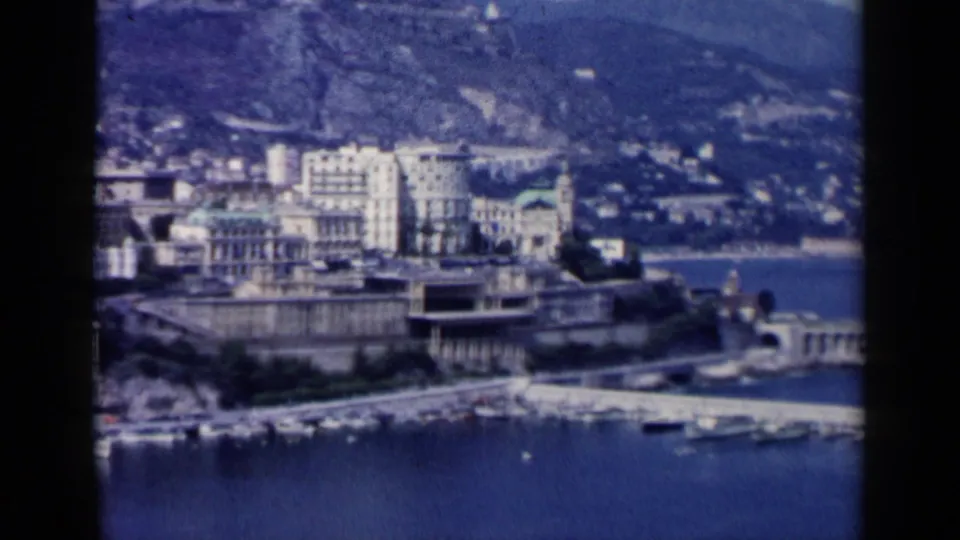 A blue-cast 1960 8mm frame shows waterside buildings, harbor frontage, and a sloping hillside behind the city.