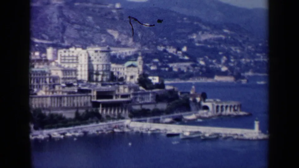 1960 travel-film frame with moored boats, a white pier or breakwater, waterfront architecture, and hills beyond.