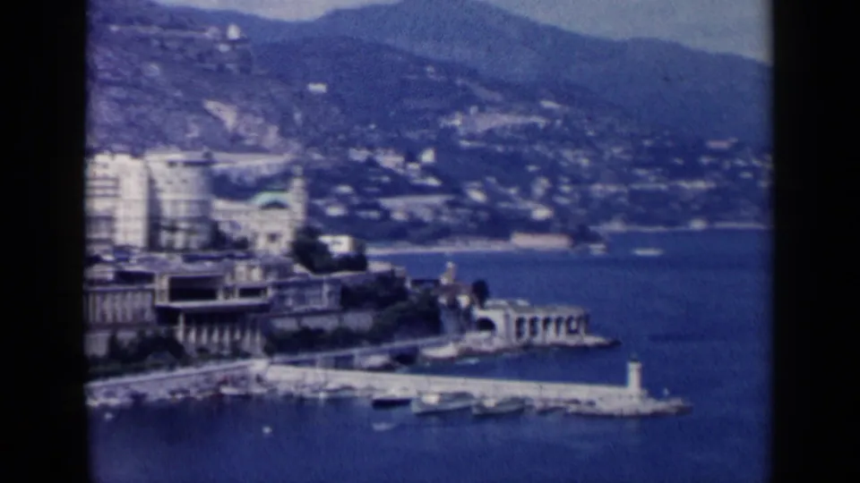 The final 1960 8mm frame looks across blue harbor water to a white breakwater, boats, waterfront buildings, and hills.