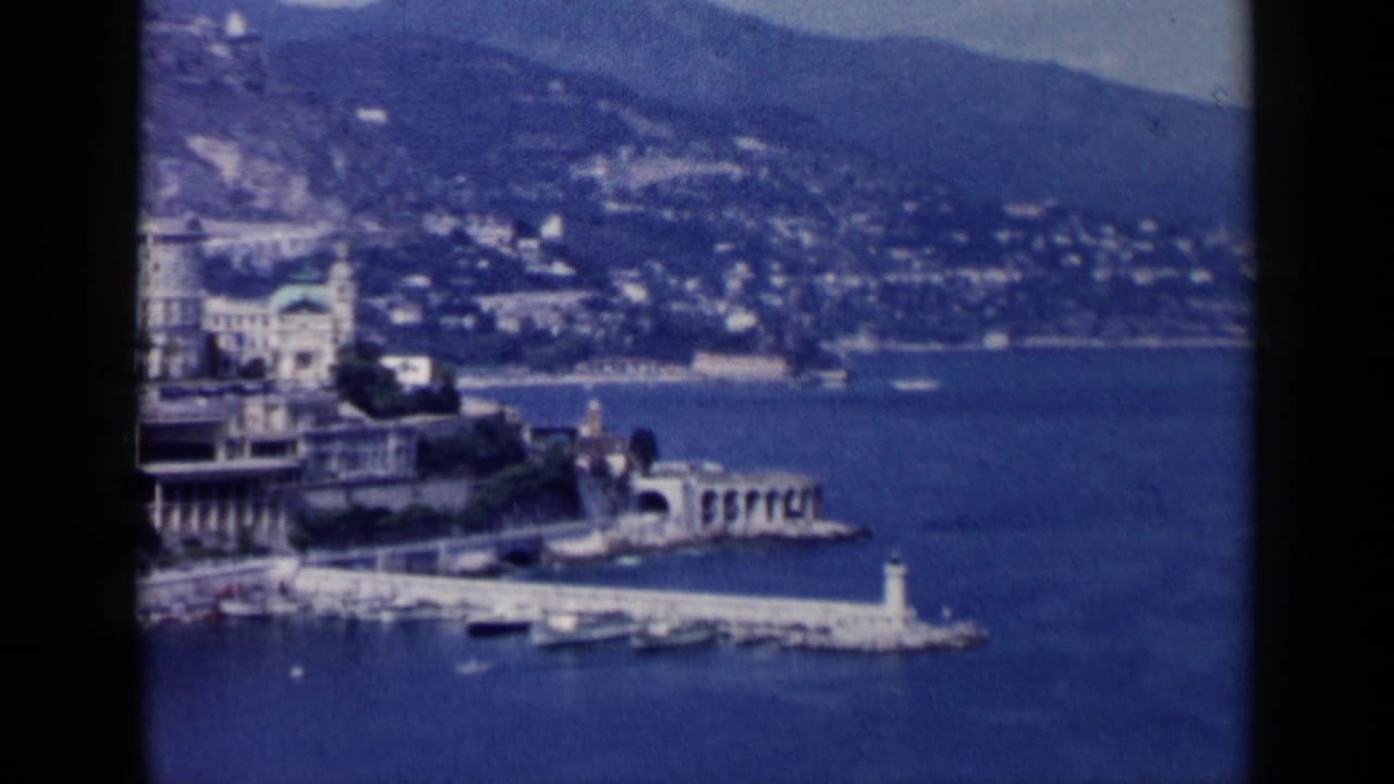 Elevated harbor view with pier, lighthouse, boats, shoreline buildings, hills, and water.