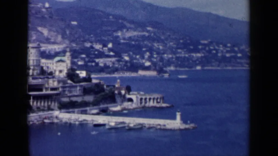 1960 8mm travel frame of a coastal harbor associated with Venice, Italy, showing a breakwater, lighthouse, boats, hillside buildings, and blue water.