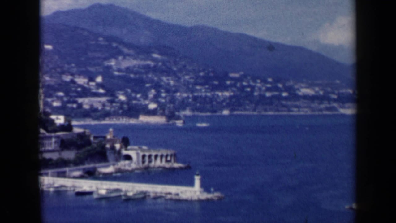 Harbor shoreline with breakwater, lighthouse, boats, hillside buildings, and mountains.