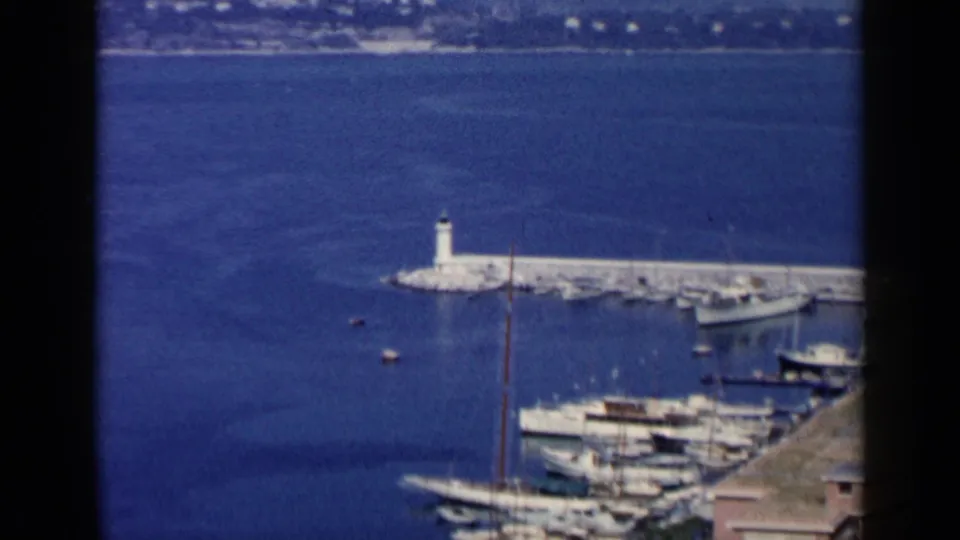 1960 home-movie frame associated with Venice, Italy, looking down at moored boats, a long pier with a lighthouse, blue water, and distant shore.