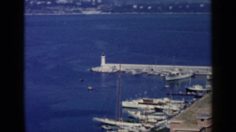 Vintage 8mm frame of the Italy harbor scene showing boats near the foreground, a lighthouse on the breakwater, open water, and distant shoreline.