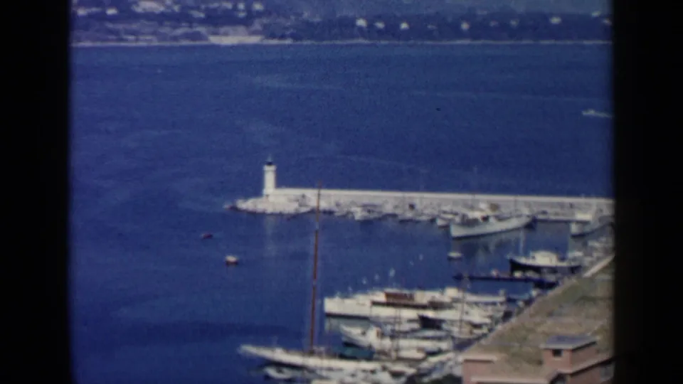 1960 8mm view of a Venice, Italy harbor with a white lighthouse on a breakwater, moored boats, blue water, and distant hillside buildings.