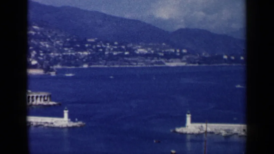 1960 Venice, Italy harbor view showing white lighthouse breakwaters set in deep blue water, backed by shoreline buildings and hills.