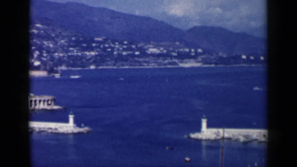 1960 8mm harbor frame from Venice, Italy with a small white lighthouse, breakwater, blue bay, hillside development, and a partial waterfront structure.