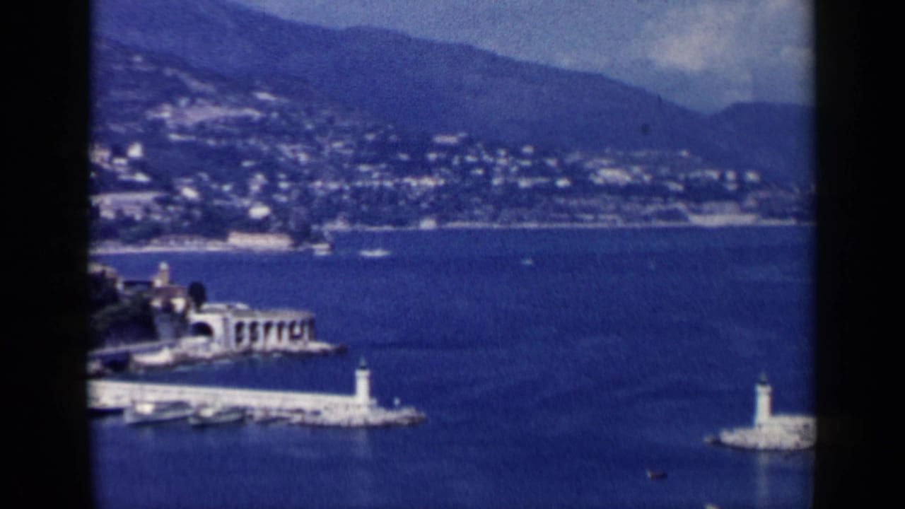1960 8mm view of Venice waterfront with harbor markers, blue water, distant hillside, and a prominent shoreline structure.