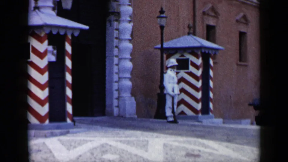 1960 Venice 8mm frame with a uniformed guard near striped booths, stonework, lamppost, and warm-toned facade.