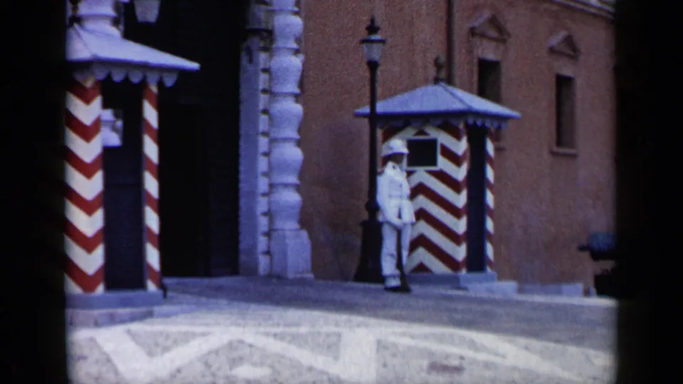 1960 Venice frame of a white-uniformed guard beside a red-and-white booth, with lamppost, stone paving, and historic wall visible.