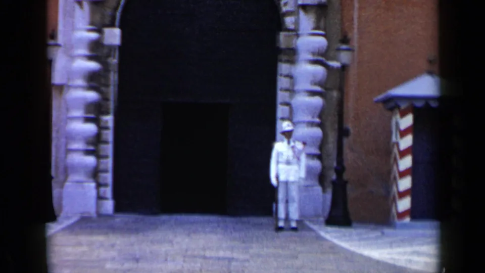 1960 Venice home-movie frame with a white-uniformed guard positioned by a dark archway, stone trim, and red-and-white sentry box.