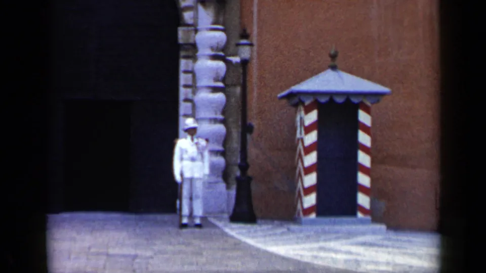 1960 Venice 8mm still with a guard at attention beside a dark arched entrance, brick facade, lamp post, and striped sentry box.