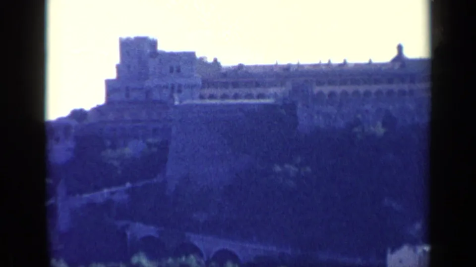Distant fortified stone structure on a high hillside in archival 1960 Italy footage, seen through soft blue-shifted 8mm film.