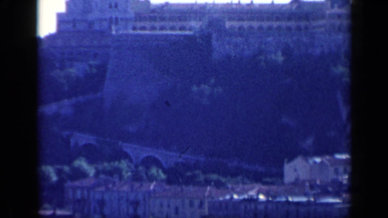 Distant blue-tinted view of a large fortified stone structure above an arched road or bridge.