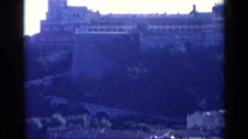 1960 Venice home-movie still showing a dark blue distant fortification, arched roadway or bridge, and lower rooflines at the frame edge.
