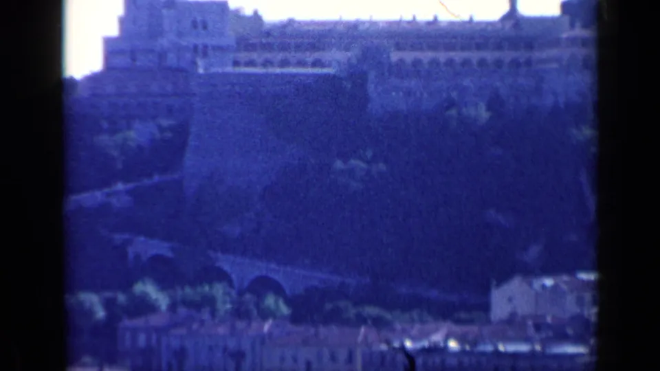 Blue-shifted 1960 Venice 8mm frame of the fortified structure, arched road or bridge, hillside planting, and small pale buildings below.