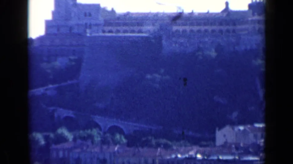Blue-tinted 1960 8mm view in Venice, Italy, showing a distant bridge, hillside, and clustered pale buildings.