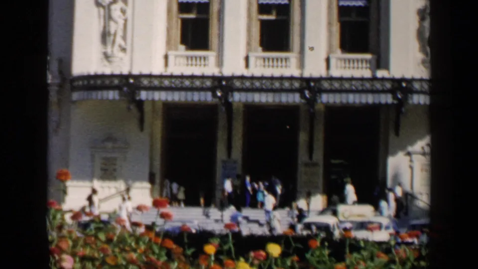 1960 Venice street view with people near a grand building entrance, parked classic cars, and bright flowers in the foreground.