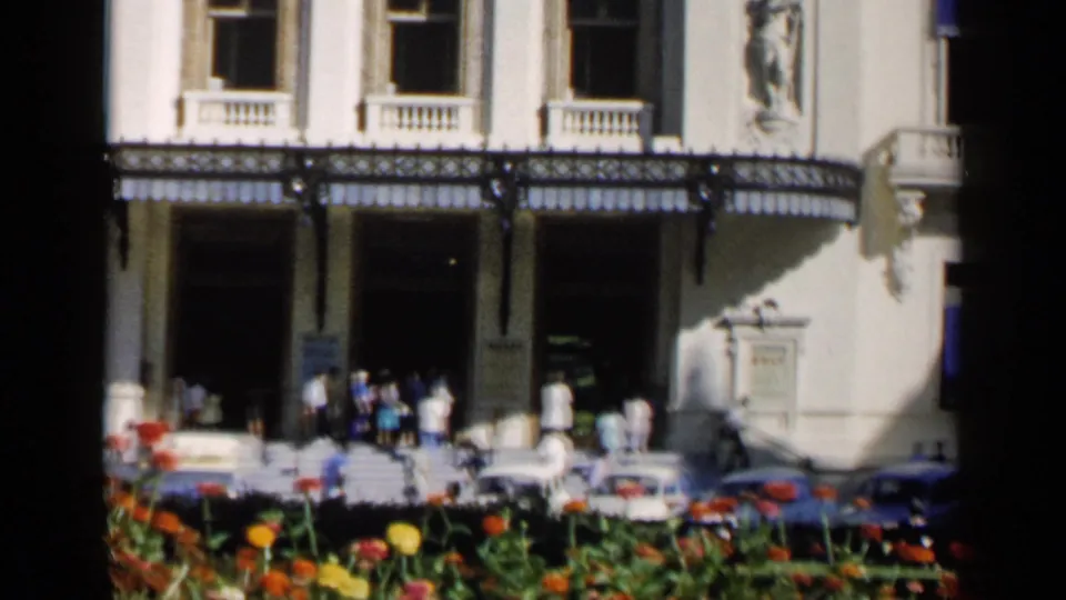 1960 Venice home-movie frame showing a grand entrance with pedestrians on the steps, parked cars below, and flowers across the foreground.