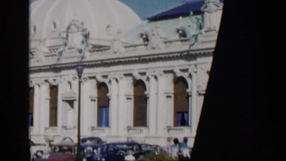 1960 Venice 8mm frame showing a pale side facade with arched windows, roofline sculpture, parked classic cars, and pedestrians below.
