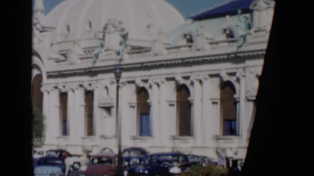 Ornate pale building facade with dome, arched windows, parked cars, and street lamps.