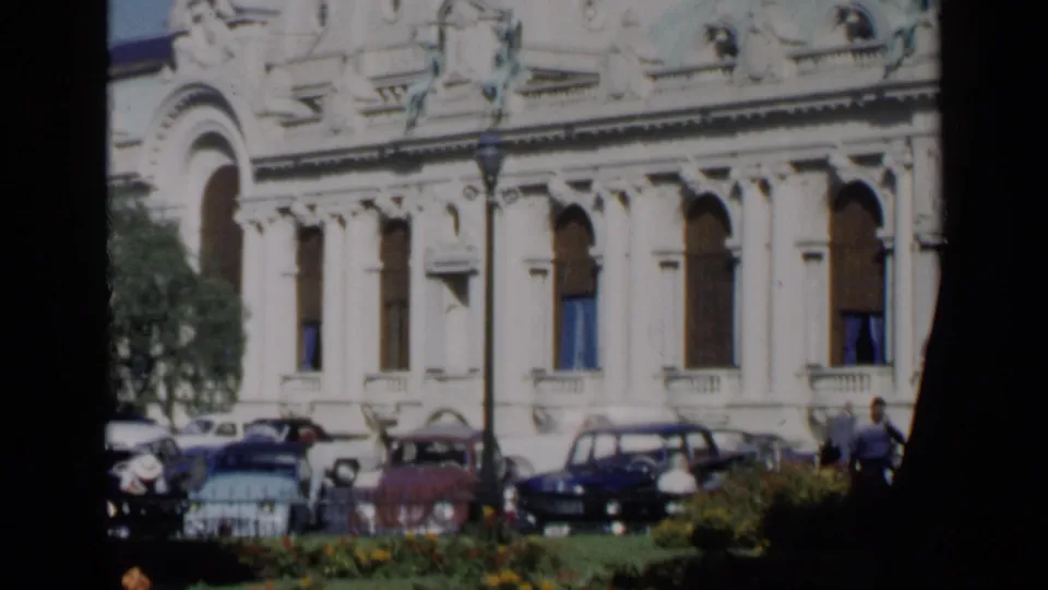 A 1960 Venice street view frames the building facade lower and closer, with arched windows, parked cars, greenery, and small pedestrian figures.