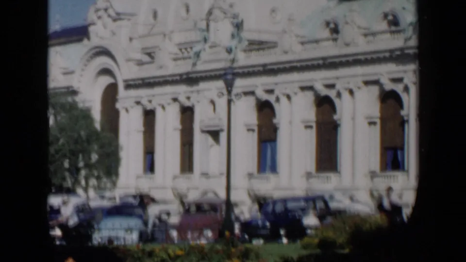 1960 Venice home-movie frame showing an ornate pale facade, arched windows, trees, and mid-century cars near the street.