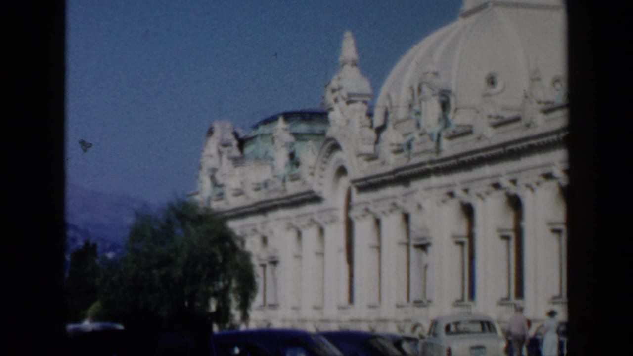 Ornate domed waterfront building with parked cars and trees under a blue sky.