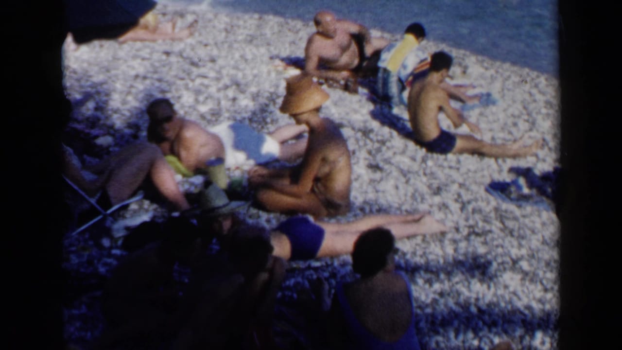 Adults in swimsuits relax on a pebble beach beside blue water.