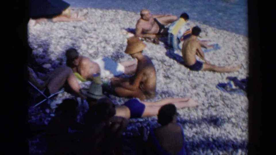 1960 Venice beach frame with adults in retro swimwear resting on pebbles, including a sun hat, towels, and water in the background.