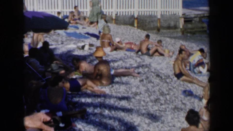 Wide 1960 Venice home-movie view of adults relaxing on a pebble beach, with a white fence, towels, umbrella shade, and blue water nearby.