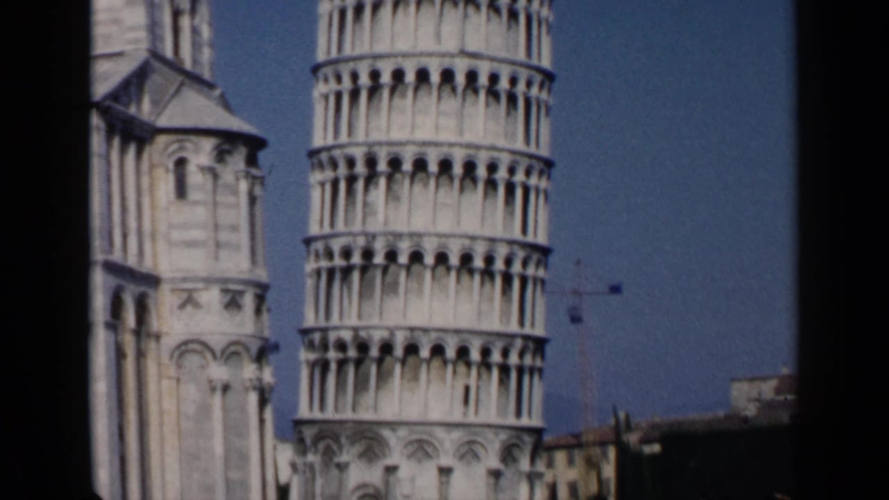 1960s 8mm view of the Leaning Tower of Pisa and neighboring cathedral structure with a clear blue sky.