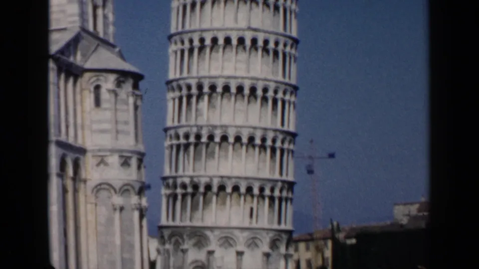 1960s 8mm view of the Leaning Tower of Pisa and neighboring cathedral structure with a clear blue sky.