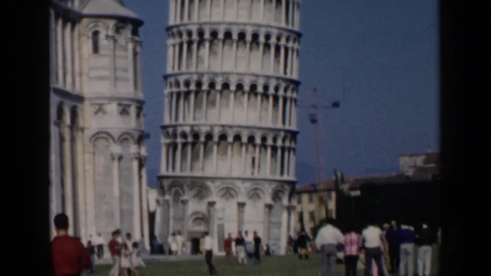1960s 8mm wide view of the Leaning Tower of Pisa, adjacent building, and tourists moving across the lawn.