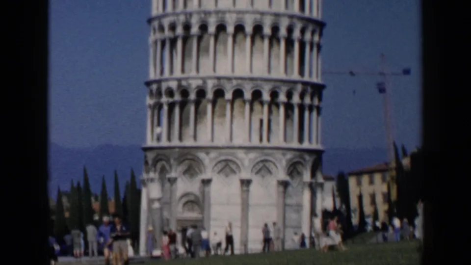 1960s 8mm view of the Leaning Tower of Pisa with visitors scattered across the grass and cypress trees in the background.