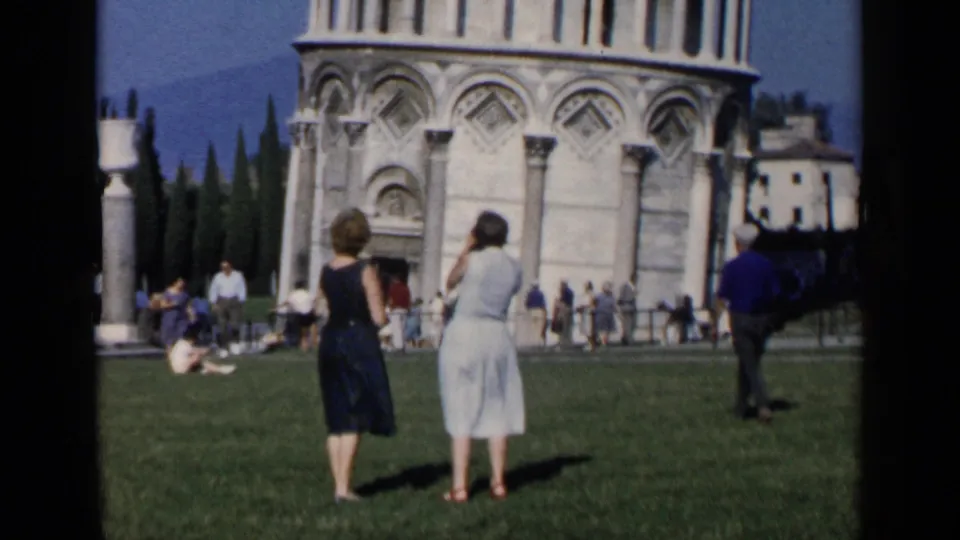 Two women in period dresses stand on the lawn near the Leaning Tower of Pisa, with tourists and cathedral architecture behind them.