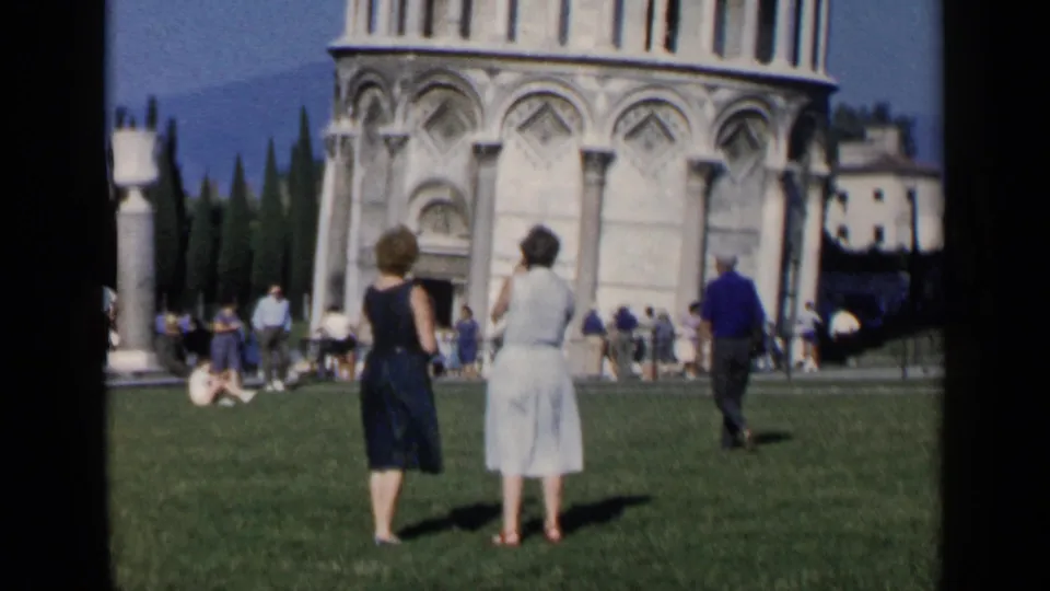Handheld 8mm frame of two women standing on the Pisa lawn, with the leaning tower base, cathedral details, and tourists in view.
