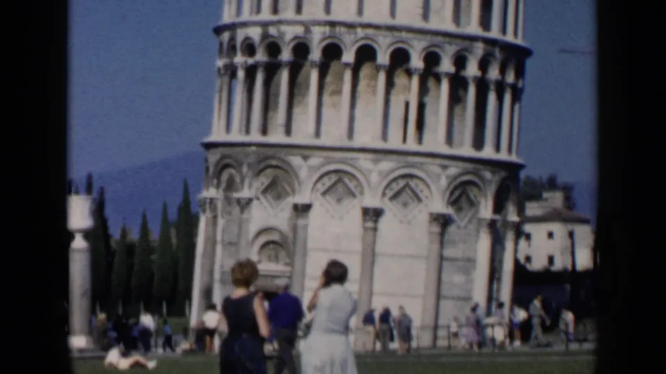 The Leaning Tower of Pisa fills the frame above tourists on the grass, with two women seen from behind in a 1960s travel-film view.