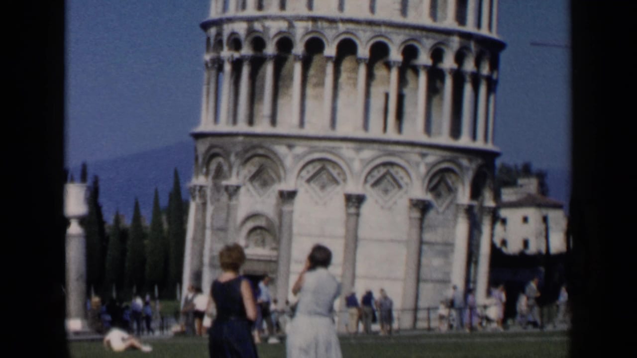 Tourists stand on grass near the base of the Leaning Tower of Pisa.