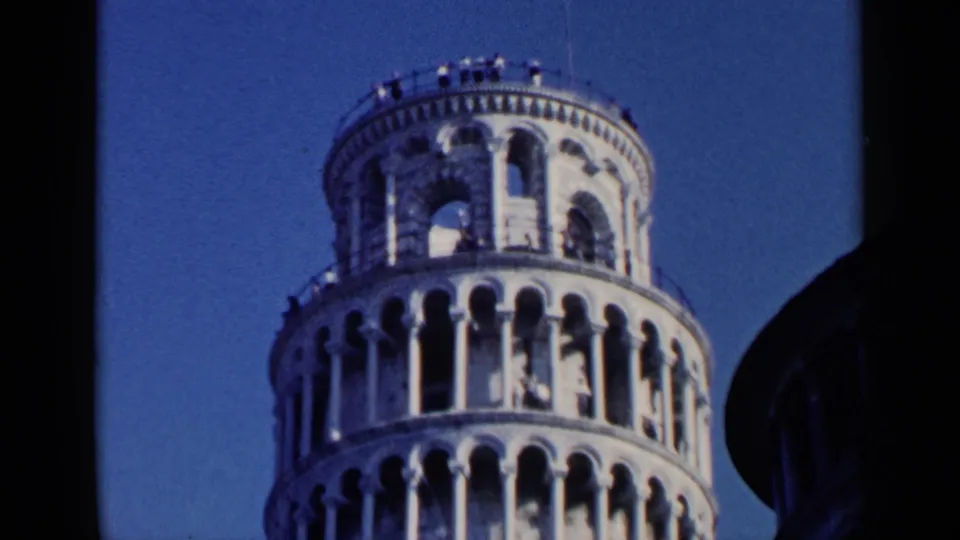 1960 8mm frame of the Leaning Tower of Pisa's upper levels, with small tourist figures visible near the railing under a blue sky.