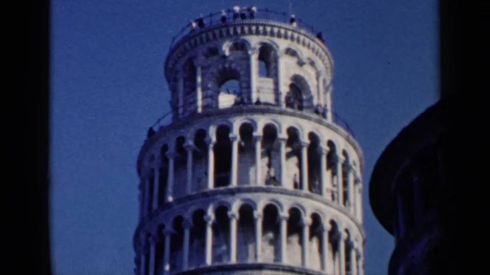 1960 8mm frame of the Leaning Tower of Pisa's upper stories, with pale stone arches, small visitors, and visible home-movie film texture.