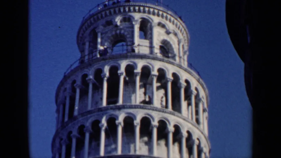 1960 8mm frame of the Leaning Tower of Pisa's upper levels, with a tourist visible near the railing under clear sky.