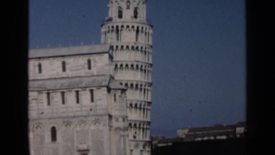 1960 8mm frame with a tighter visitor view of the Leaning Tower of Pisa, the cathedral wall, and a dark border at the edge.