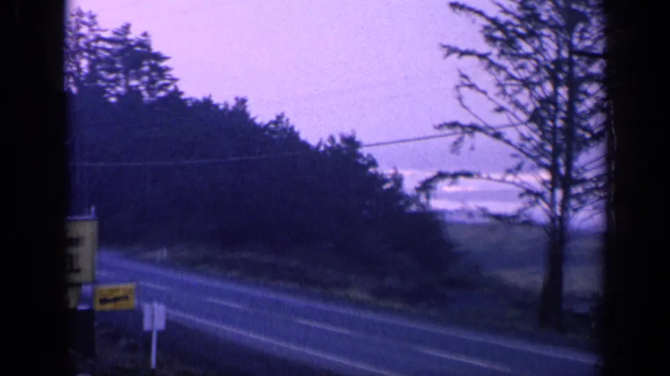 1963 California home-movie frame with the motel sign nearly out of view beside a quiet tree-lined highway.