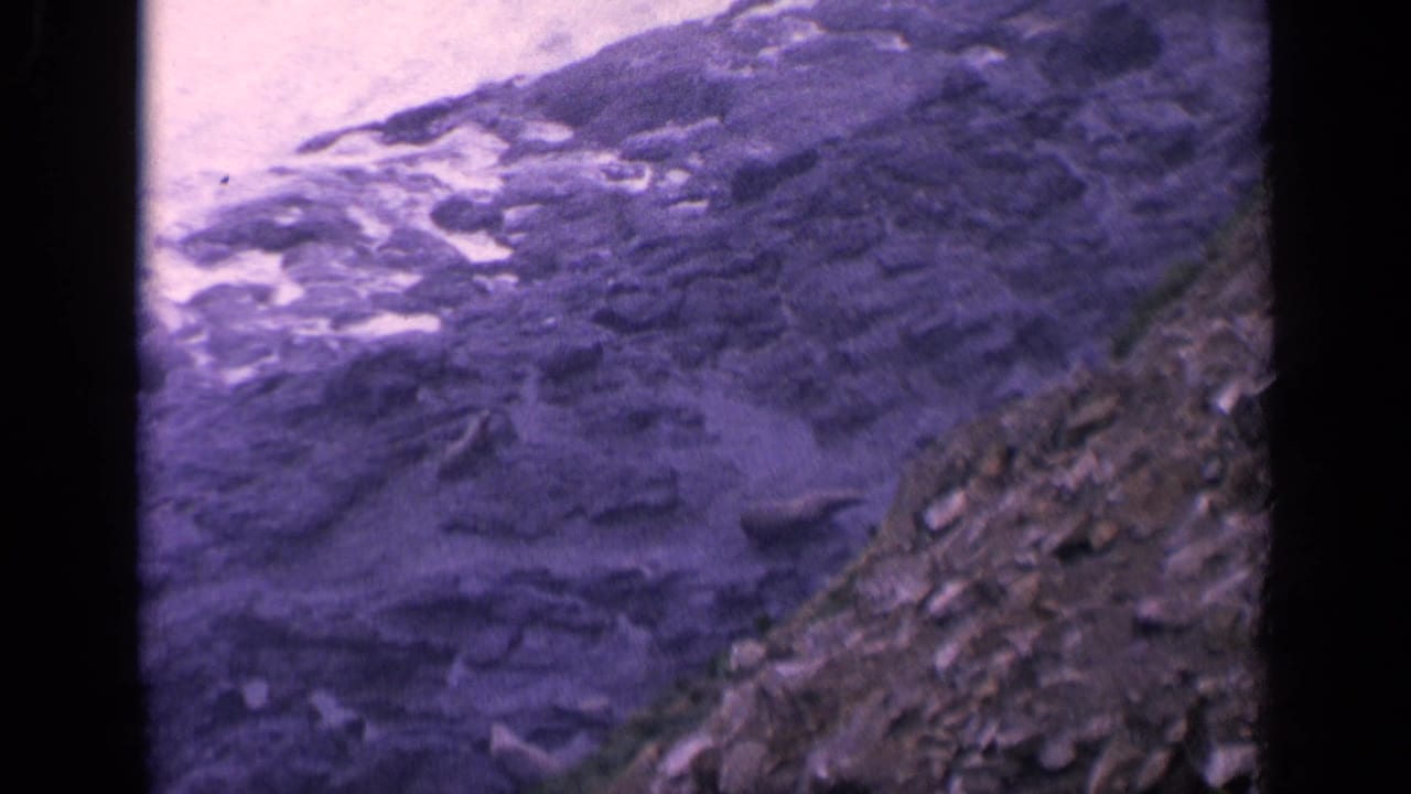 Waves wash over dark layered rocks beside a sloped rocky shoreline.